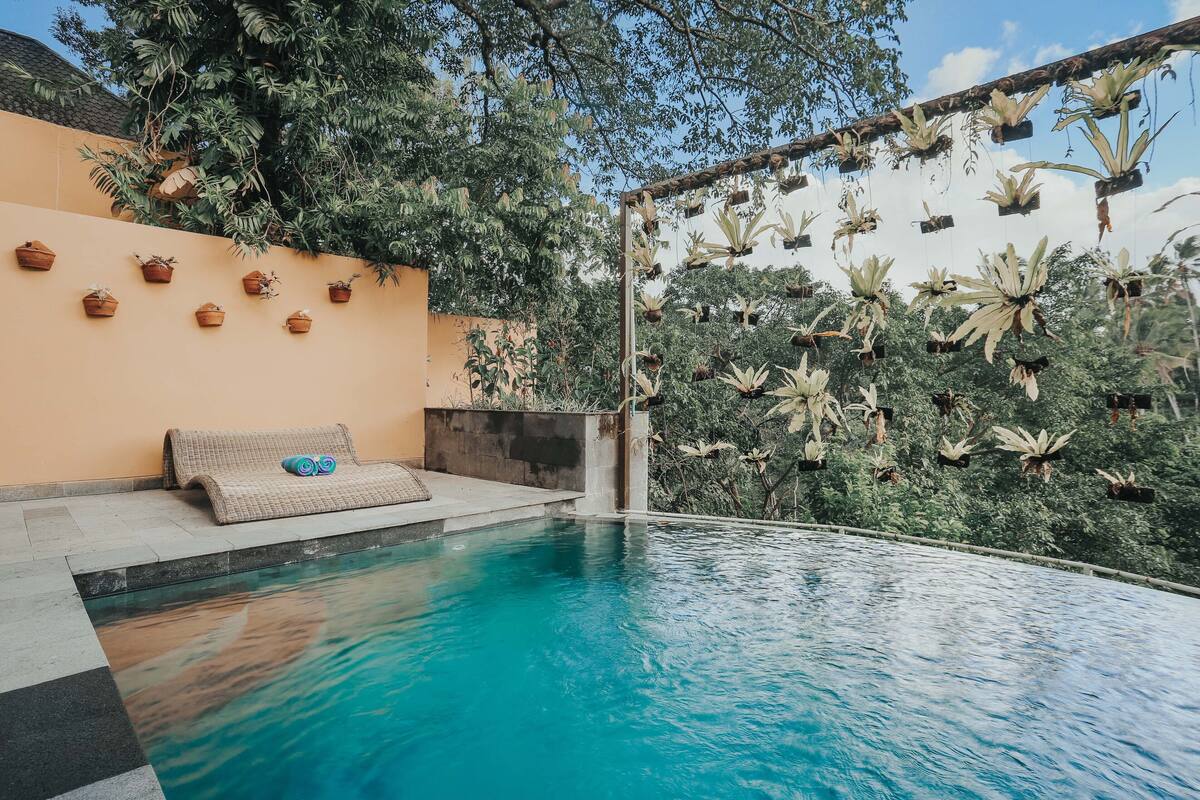 An infinity pool with a clear blue water surface, surrounded by a low wall with potted plants and a wicker bench.