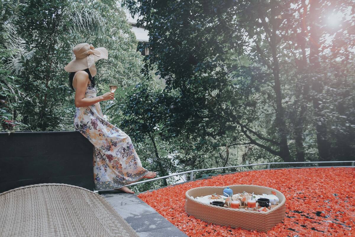 A woman in a floral dress and wide-brimmed hat standing on a balcony with a wicker basket containing drinks and snacks.