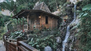 Una casa de madera con techo de tejas, rodeada de abundante vegetación y una pequeña cascada.