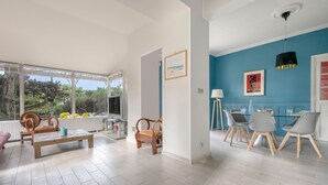 Un vaste espace de vie comprenant une table basse en bois, deux chaises en bois, une table à manger avec quatre chaises, et une vue sur l’extérieur grâce à de grandes fenêtres.