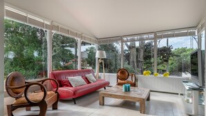 Un salon spacieux avec un canapé rouge, des chaises en bois, une table basse et un téléviseur. De grandes fenêtres offrent une vue sur la verdure environnante.
