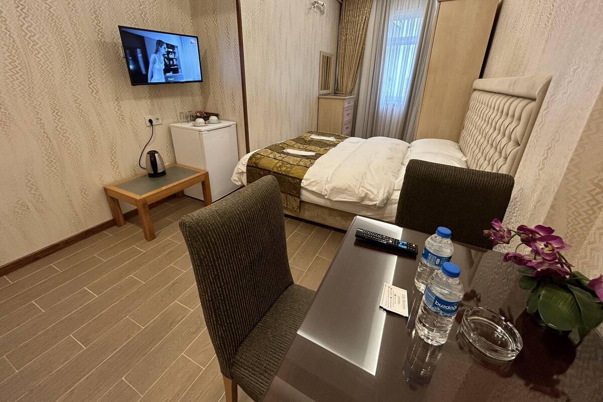 A hotel room with a bed, a desk with a chair, a TV mounted on the wall, a small table with a kettle, a water bottle, and a vase with flowers.