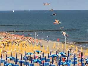 A beach with rows of blue and yellow umbrellas, a sandy shore, and a view of the sea with several kites flying.