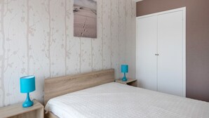 Un dormitorio con cabecera de madera, colcha blanca, dos mesitas de noche con lámparas, un cuadro en la pared y un armario.