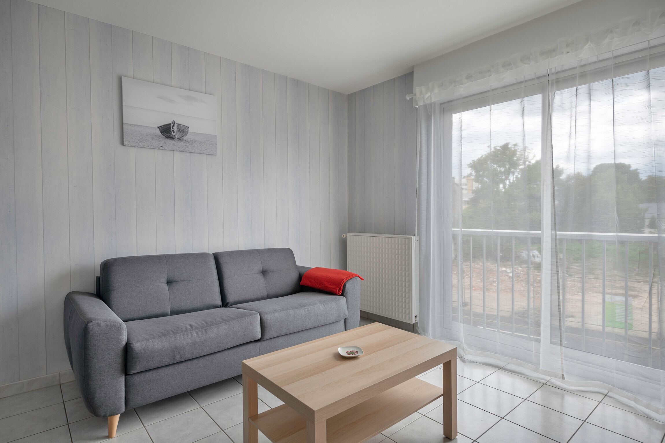 A living room with a grey sofa, a wooden coffee table, and a large window with sheer curtains.