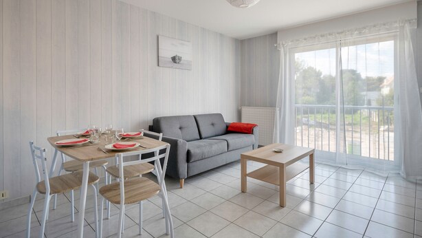 Una sala de estar moderna con un sofá gris, paredes blancas y una mesa de comedor preparada para cuatro comensales.