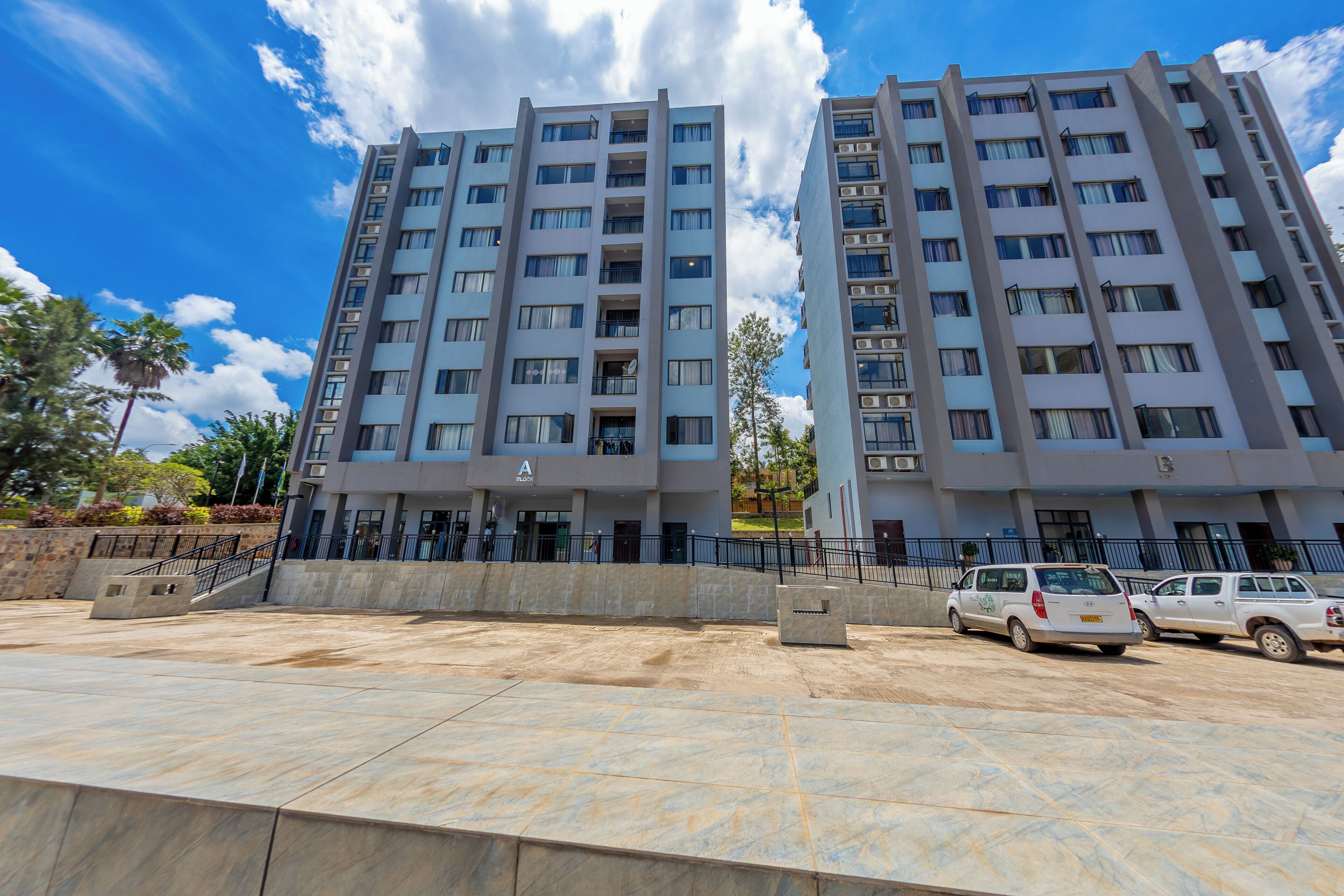 A modern multi-story residential building with a flat facade, balconies, and a parking area with vehicles.