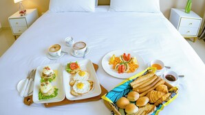 Ein ordentlich bezogenes Bett mit weißen Leinen, ein Frühstücksbrett mit Eiern, Toast und Obst sowie ein Korb mit Brötchen in einem Hotelzimmer.