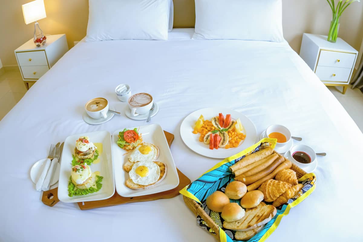 A neatly made bed with white linens, a breakfast tray with eggs, toast, and fruit, and a basket of rolls on a hotel room.