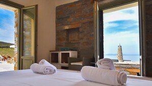 A hotel room with a bed, two rolled towels, a stone fireplace, and a view of the sea and a lighthouse. - Villa LENA:Beachfront Villa 70 m to sandy isolated Beaches C/spa outdoor jacuzzi (Kea)