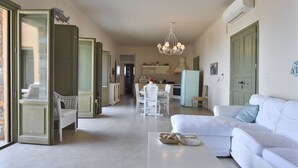 A spacious living room with a white sofa, a dining area with a white table and chairs, and a kitchen in the background. - Villa LENA:Beachfront Villa 70 m to sandy isolated Beaches C/spa outdoor jacuzzi (Kea)
