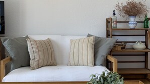 A cozy living room with a sofa, cushions, a wall-mounted light fixture, a shelf with decorative items, and a woven basket with a plant. - CANYELLES PETITES - ROSAS 3mn plage Vue mer Tout confort GARAGE Terrasse 40m2 (Roses)