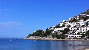 A beach with white buildings on a hillside, clear blue water, and a sandy shore. - CANYELLES PETITES - ROSAS 3mn plage Vue mer Tout confort GARAGE Terrasse 40m2 (Roses)