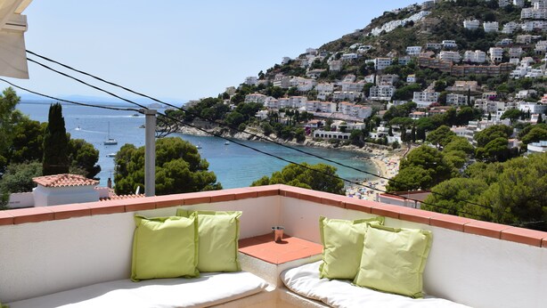 A balcony with white cushions and green pillows, overlooking a coastal town with buildings on a hillside and boats on the water. - CANYELLES PETITES - ROSAS 3mn plage Vue mer Tout confort GARAGE Terrasse 40m2 (Roses)