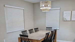 Een eetgedeelte met een houten tafel en zwarte stoelen, witte zonwering aan de ramen en een hangende lamp.