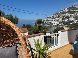 A balcony with a wicker chair, a potted plant, and a view of a coastal town with buildings and a body of water.