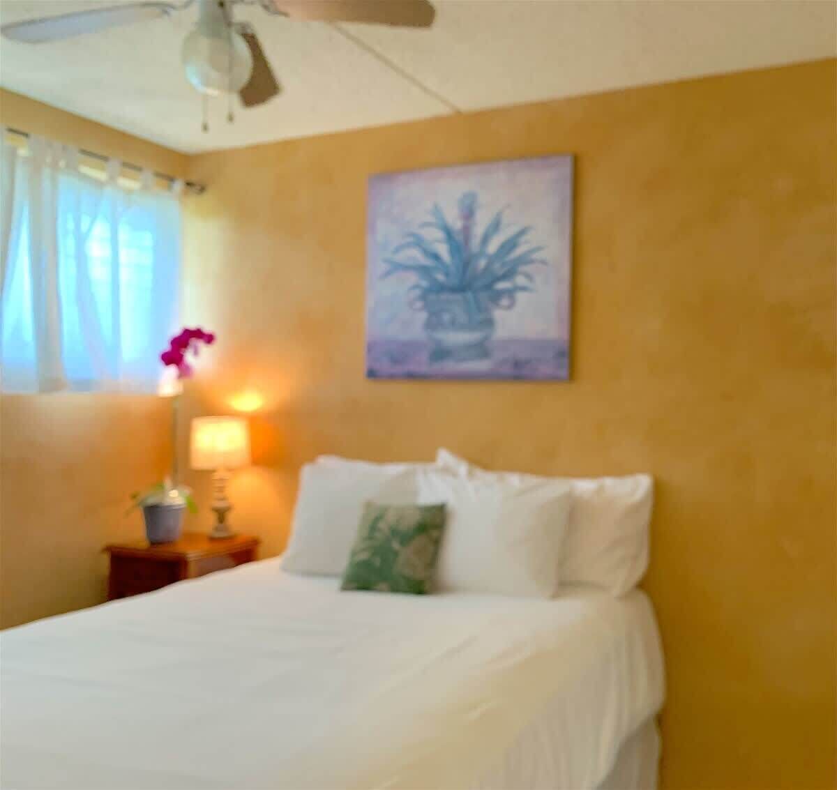A bedroom with a bed, a ceiling fan, a window with curtains, a nightstand with a lamp, and a wall painting.