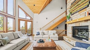 A spacious living room with a high ceiling, large windows, a fireplace, and a staircase leading to an upper level. - Western Sky Ranch | 8 Bed | Pool Table + Deck (Breckenridge)