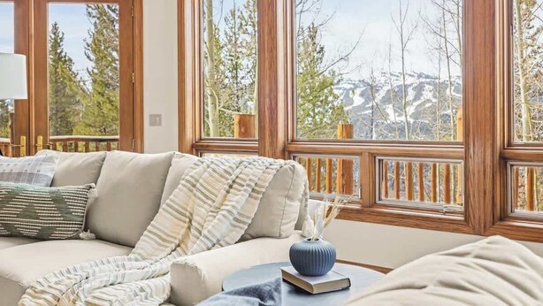 A cozy living room with a beige sofa, a striped throw blanket, and a small table with a vase and book. Large windows offer a view of trees and mountains. - Western Sky Ranch | 8 Bed | Pool Table + Deck (Breckenridge)