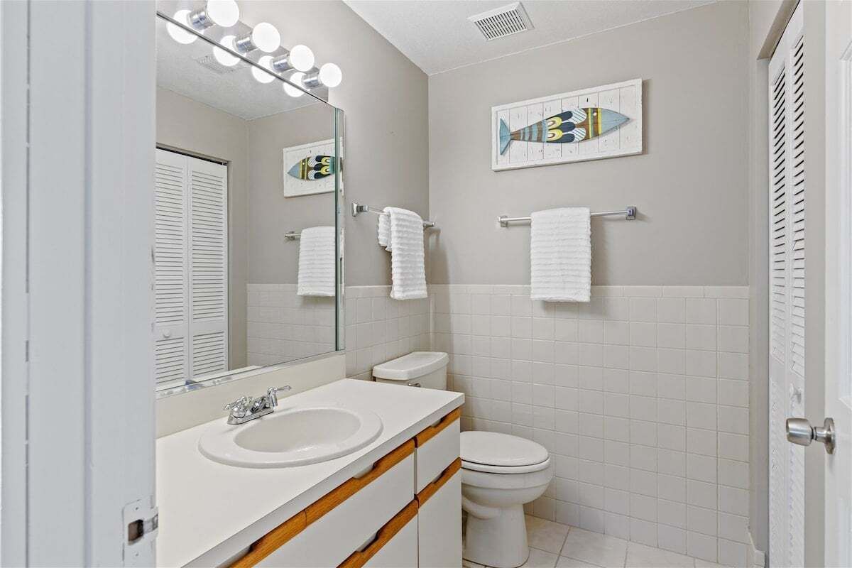 A bathroom with a double sink, a toilet, and a mirror with lights. There are towels on a rack and a fish-themed artwork on the wall.
