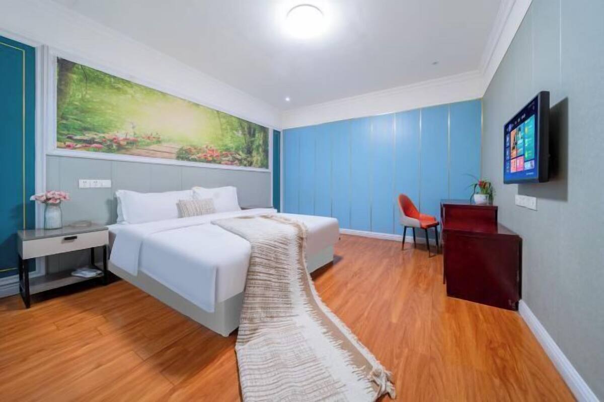 A bedroom with a bed, a nightstand, a chair, a desk, a painting, and a TV on the wall.