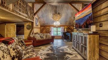 A rustic cabin interior with a loft bedroom, a dining area, and a living room furnished with a red sofa and a cowhide rug.