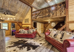 A rustic interior with wooden walls, a wooden staircase, two leather sofas with patterned cushions, a cowhide rug, and a digital clock on the wall.