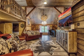 Een rustiek interieur met een houten plafond, een bank met kussen van runderleer, een vloerkleed van runderleer en een schilderij van een berglandschap.