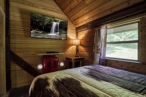 A wooden cabin room with a bed, a mounted TV displaying a waterfall, a table with a lamp, and a window with curtains.