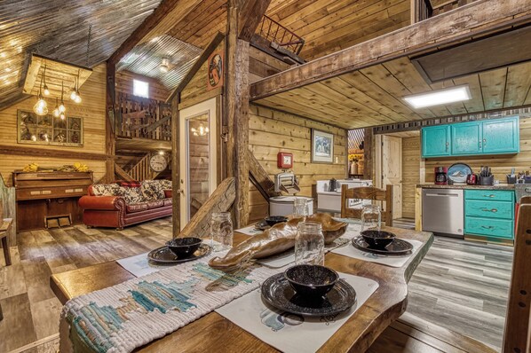 A rustic interior with a wooden dining table set for four, a sofa, a kitchen area with teal cabinets, and a staircase leading to an upper level.