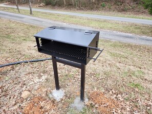 A portable barbecue grill with a metal frame and a flat cooking surface, set on a grassy area with a road in the background.