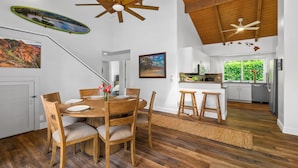 A spacious dining area with a wooden table and chairs, a ceiling fan, and a view of the kitchen and outdoor greenery. - Laule'a 'Ohana Hale - Luxurious 'Ohana home with A/C (Princeville)