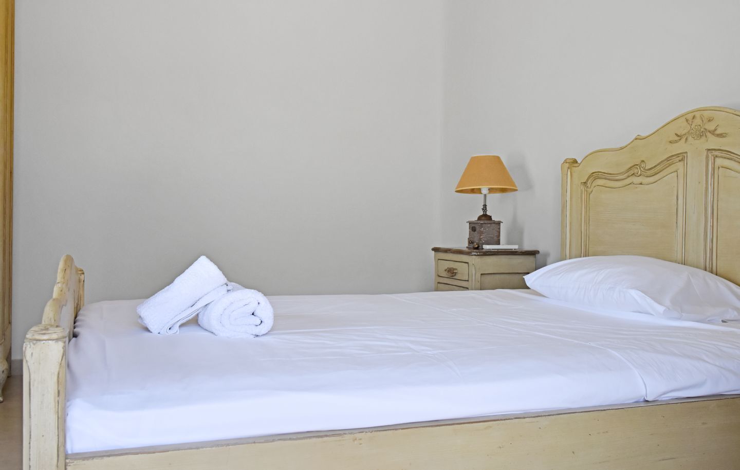 A bed with white linens, a rolled towel, a bedside table with a lamp, and a nightstand with a drawer.