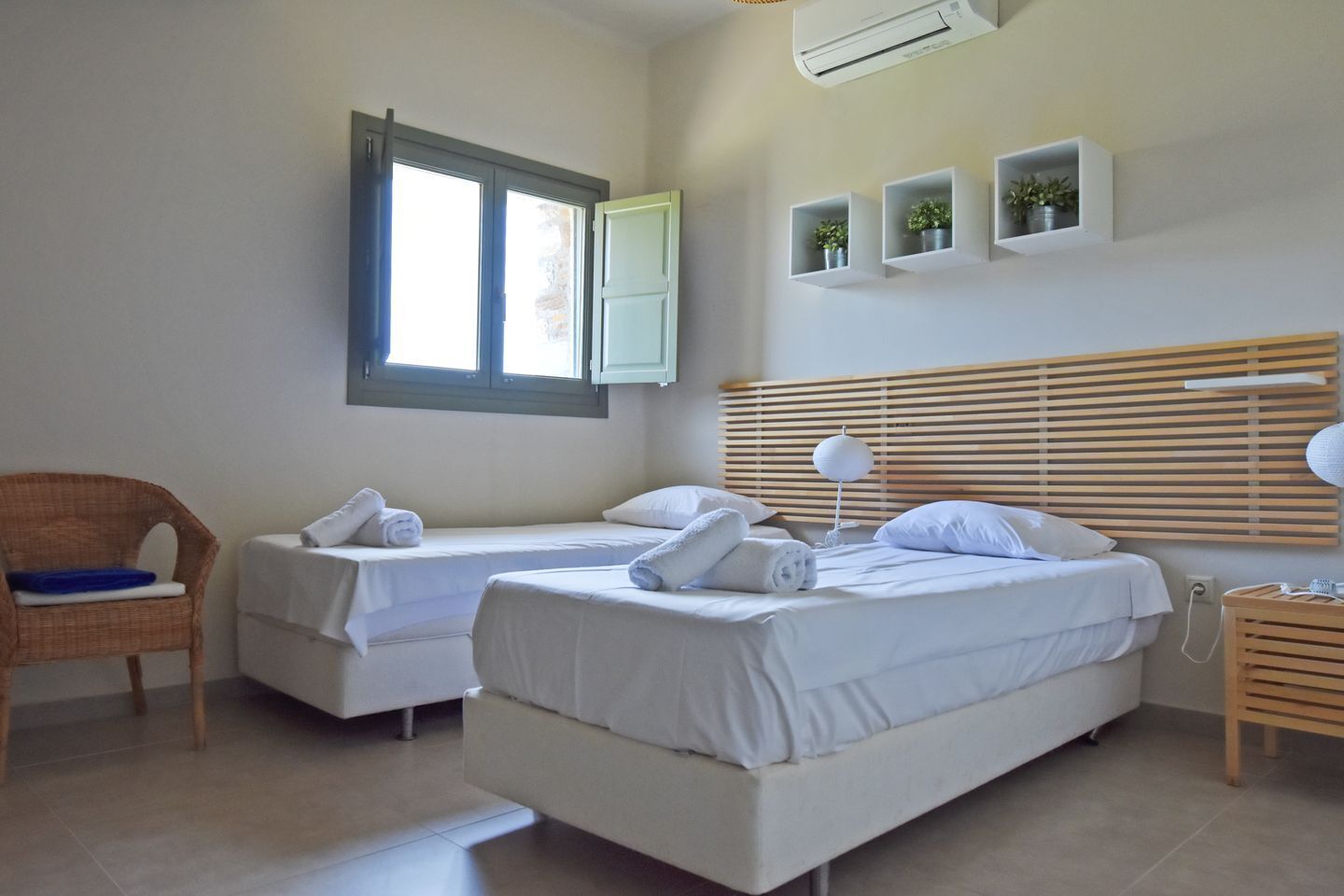A hotel room with two single beds, a wicker chair, a bedside table with a lamp, and a window with blinds.