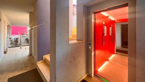 Un couloir avec un ascenseur rouge, un escalier et une porte donnant sur une pièce dont un mur est rose.