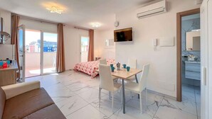 A modern hotel room with a bed, a dining area with a table and chairs, a TV mounted on the wall, and an air conditioning unit.