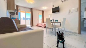 A modern living room with a sofa, a dining table with chairs, a TV mounted on the wall, and a dog.
