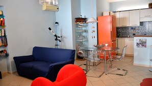 Ein gemütlicher Wohnraum mit einem blauen Sofa, einem Glastisch mit Stühlen, einer Küchenzeile mit weißen Schränken und einer Bücherwand voller Bücher.