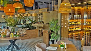 Interior moderno de una cafetería con una mesa y sillas de madera, lámparas naranjas colgantes y una estantería para vinos al fondo.