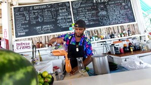 Una persona preparando una bebida en un bar, con un panel de menú detrás que muestra diferentes bebidas y platillos.