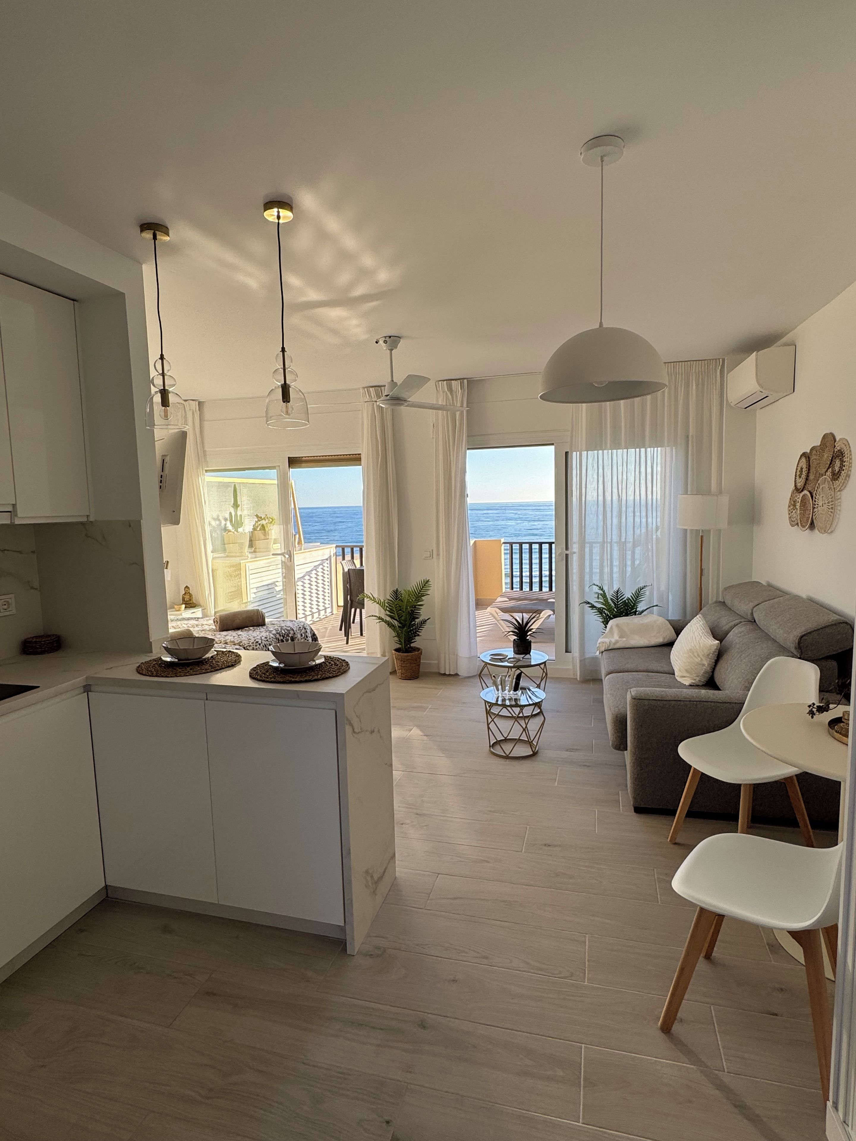 A modern kitchen with a central island, a dining area with a table and chairs, a sofa, and a view of the sea.