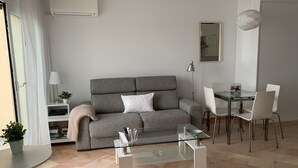 En moderne stue med en grå sofa, et glasbord, et spiseområde med et glasbord og hvide stole samt et lille bord med lampe og en potteplante.