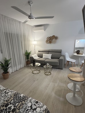 A modern living room with a grey sofa, two side tables, a ceiling fan, and a dining area with white chairs and a small table.