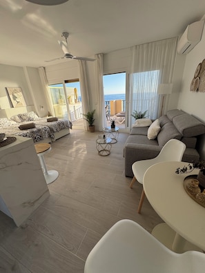 A modern living room with a sofa, a dining table, and a balcony with a view of the sea.