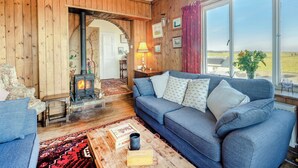 A cozy living room with a blue sofa, a wooden fireplace, and a large window offering a view of the countryside. - Coastguard's Cottage (Weybourne)