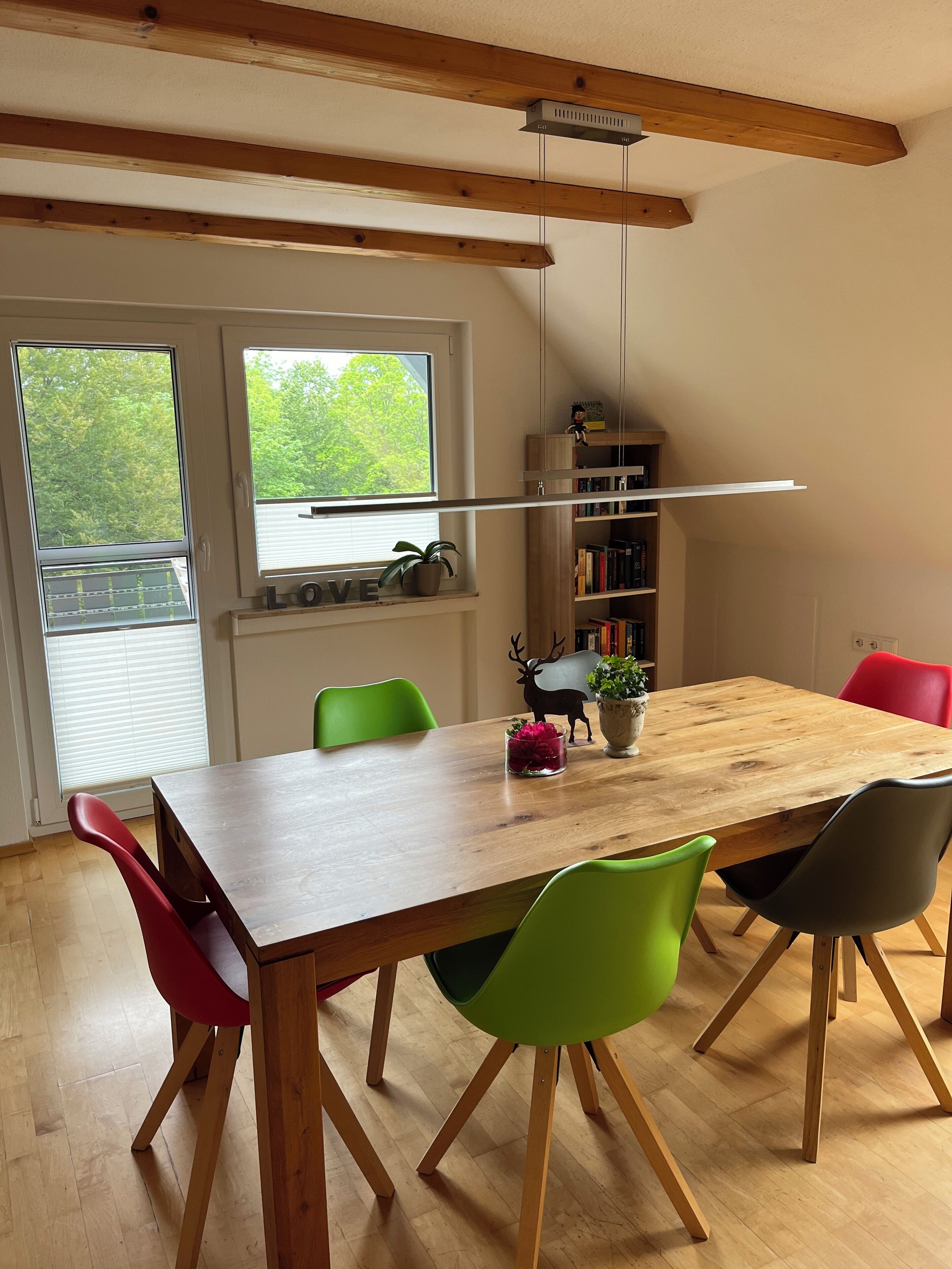 A dining area with a wooden table, green and red chairs, a bookshelf, and a window with a view of trees.