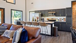 Cocina moderna con gabinetes oscuros, un lavamanos de acero inoxidable y una isla grande. En primer plano, un sofá de cuero con cojines estampados.