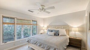 Ein Schlafzimmer mit Bett, Nachttisch, Lampe, Deckenventilator und großen Fenstern mit Jalousien.