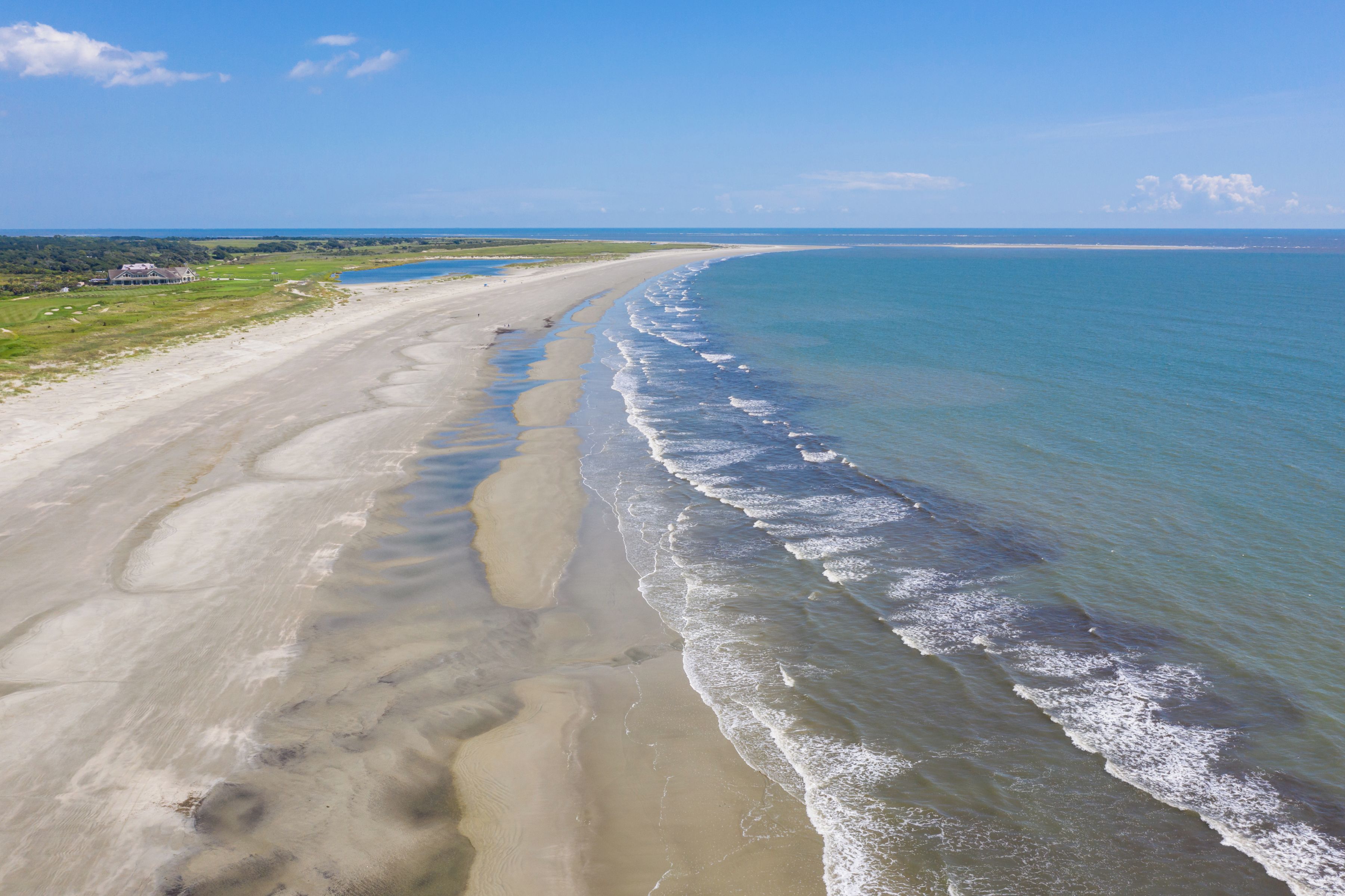 A beach with a long stretch of sand and gentle waves, a clear sky, and a distant shoreline.