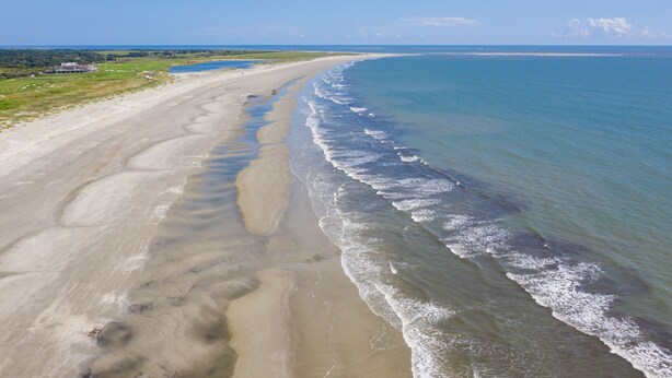 Ein Strand mit einer weiten Sandfläche und sanften Wellen, einem klaren Himmel und einer fernen Küstenlinie.